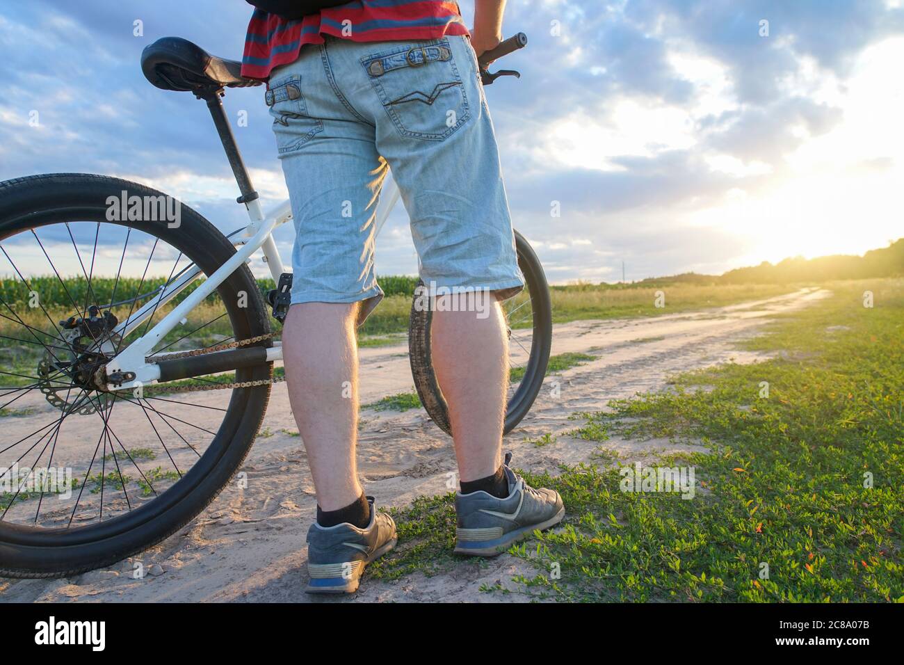 Un ragazzo su una mountain bike in una T-shirt rossa e con uno zaino corre su una strada sterrata al tramonto Foto Stock