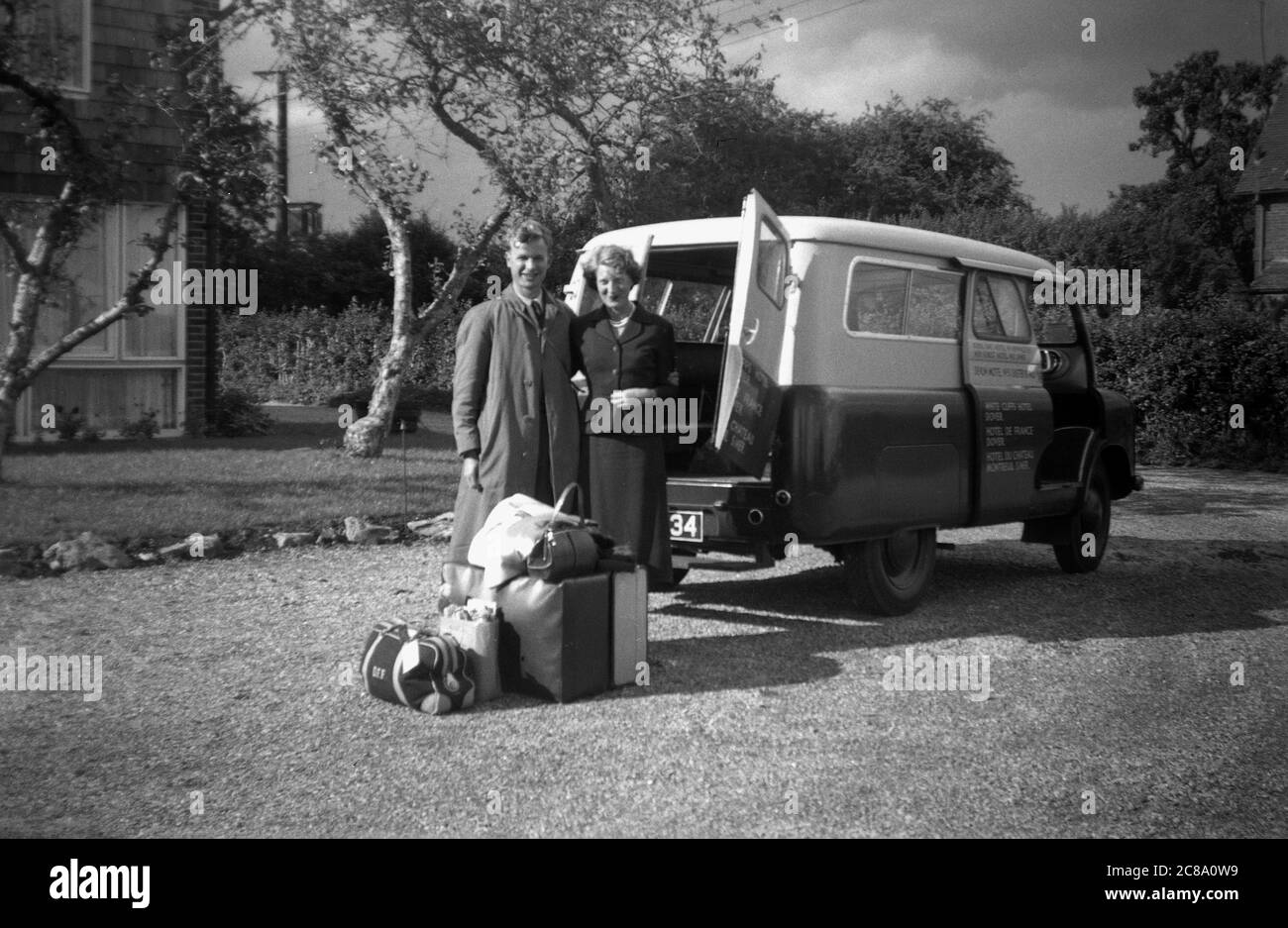 Anni '50, storico, una coppia in piedi con i loro bagagli, in attesa su un vialetto di trasporto dal loro, un piccolo pulmino, forse un van Bedford, con una porta aperta sul retro, fuori il White Cliffs Hotel, dover, Kent, Inghilterra, prima di andare a prendere il traghetto per la Francia. Foto Stock