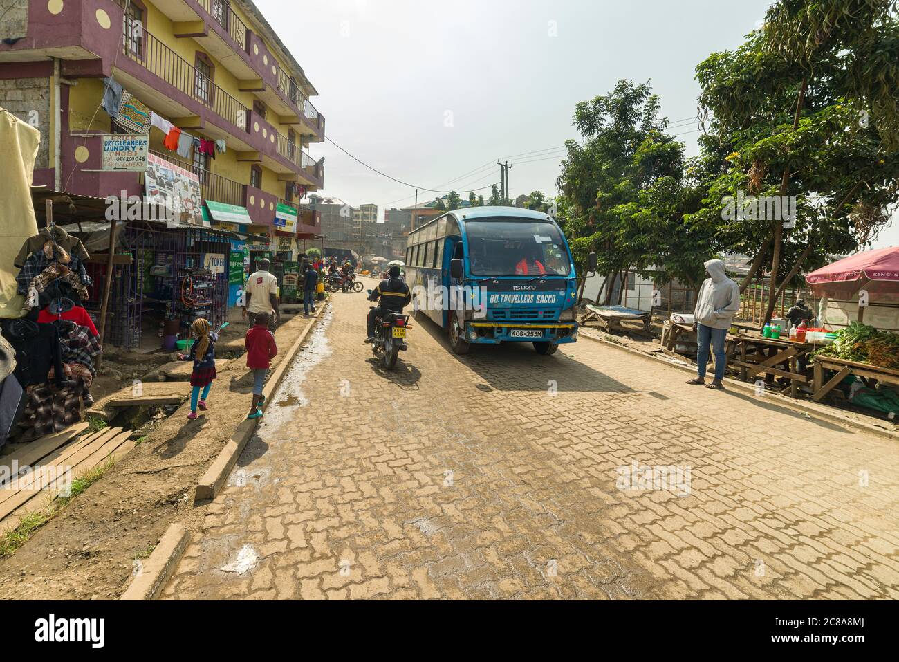 Un autobus e moto guidano giù una strada lastricata con gli edifici e la gente da una parte, Korogocho, Nairobi, Kenya Foto Stock