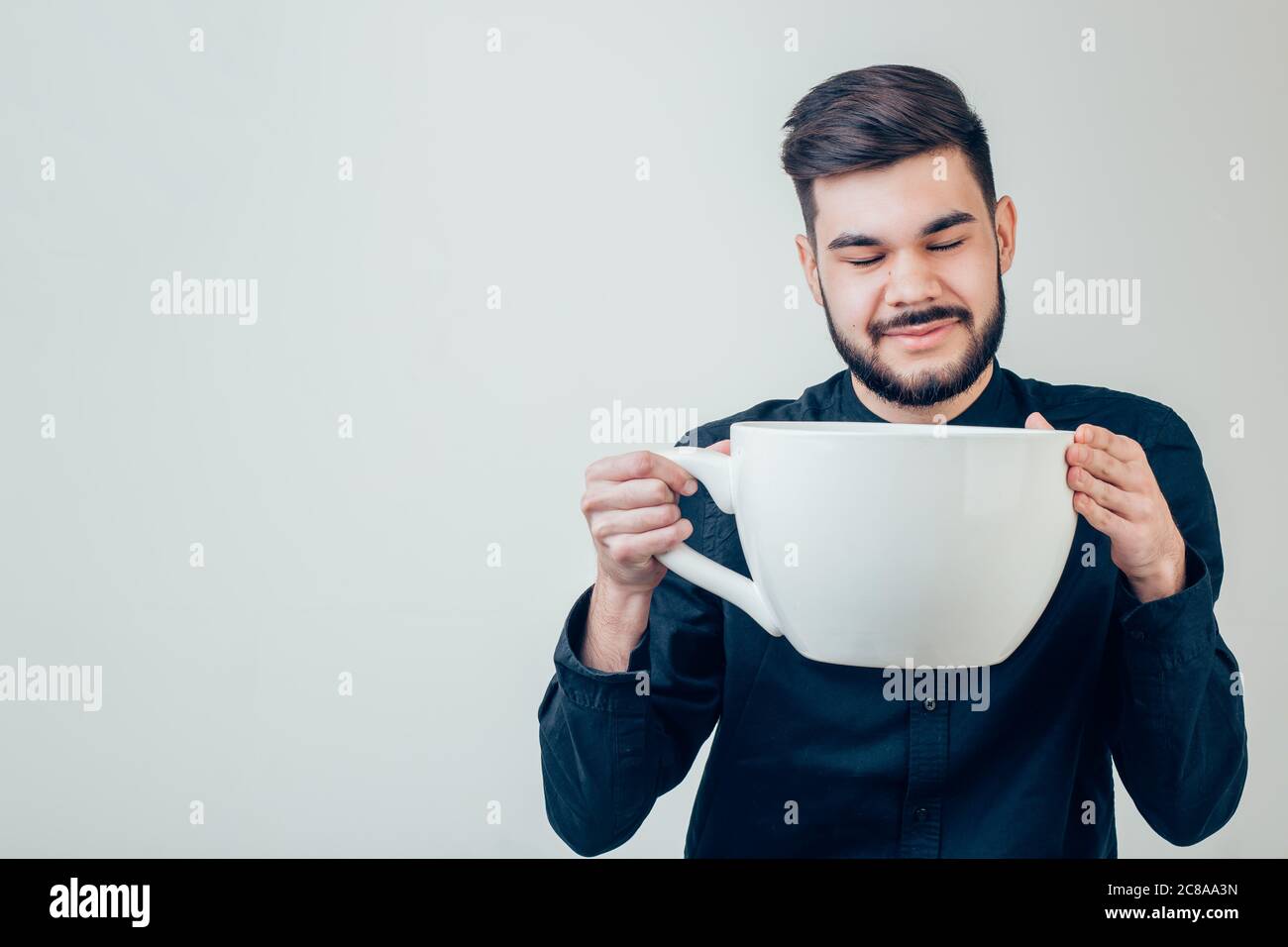 giovane uomo d'affari felice che tiene una tazza enorme e divertente di caffè nero oversize in concetto di dipendenza da caffeina isolato su sfondo uniforme Foto Stock