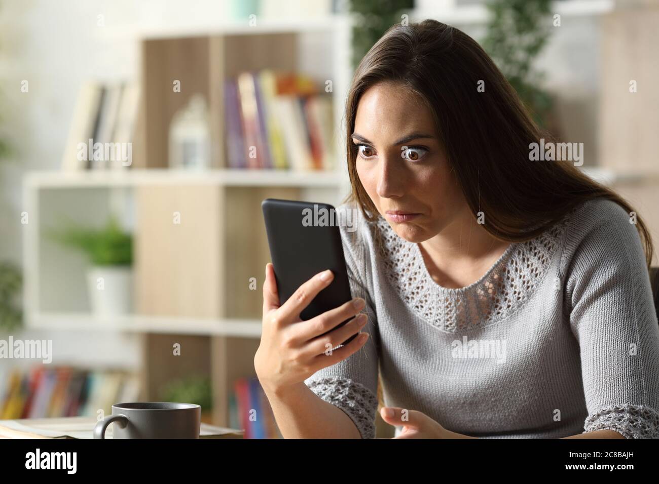 Donna perplessa che guarda sorpreso di smartphone seduto su una scrivania a casa Foto Stock