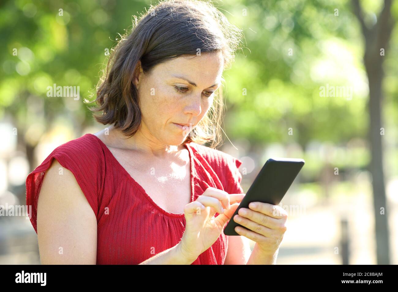 Donna adulta seria che usa lo smartphone in piedi in un parco una giornata di sole Foto Stock