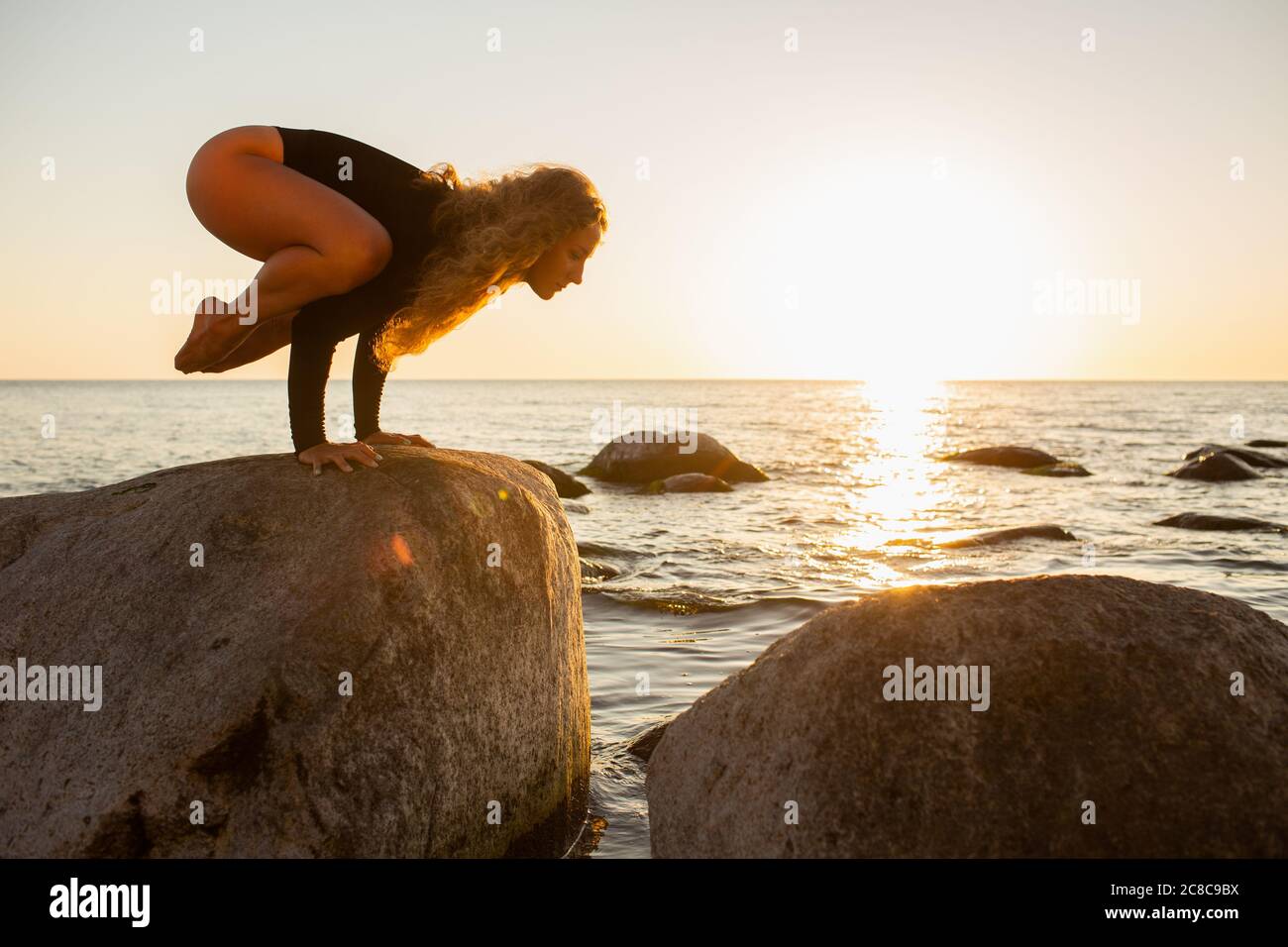 Silhouette yoga ragazza sulla spiaggia all'alba facendo Crane posa. Ragazza dai capelli lunghi che pratica yoga su una grande pietra. Bakasana Foto Stock