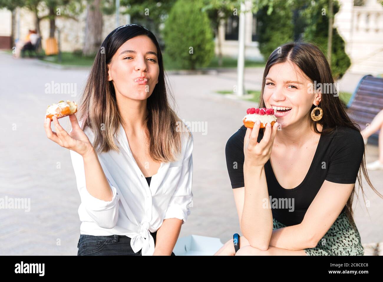 due ragazze attraenti e allegre che si godono deliziose ciambelle nel parco cittadino in un umore positivo ed energico Foto Stock