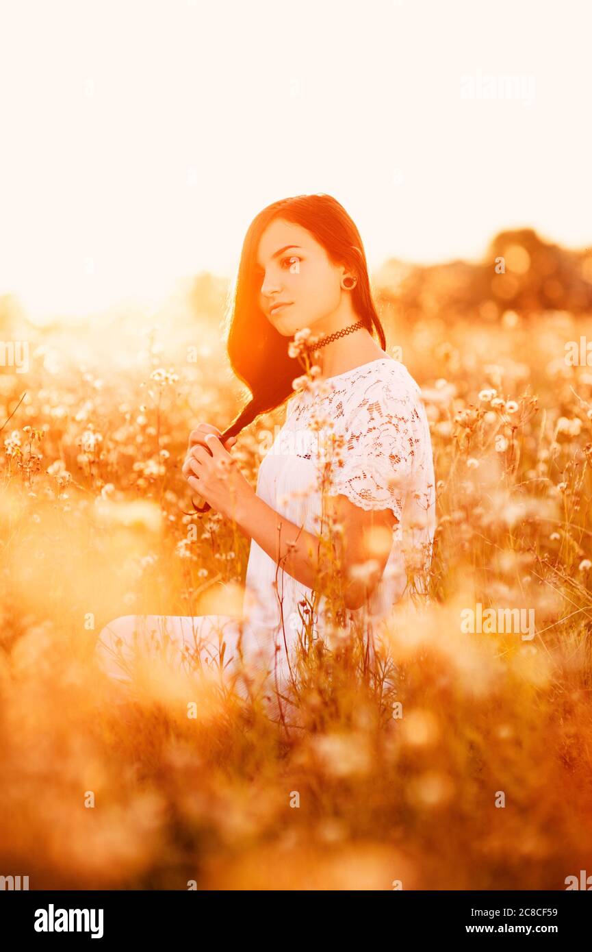 Bella tenera giovane donna generazione Z con capelli lunghi in un campo di fiori in estate in abito al tramonto sullo sfondo, stile di vita sano concetto di libertà Foto Stock