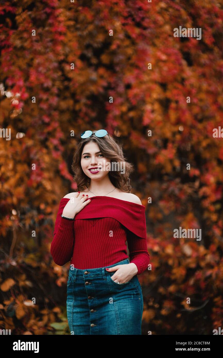 Bella giovane donna casual in occhiali che si trova tra giallo e rosso foglie nel parco pubblico, colori autunno sfondo Foto Stock