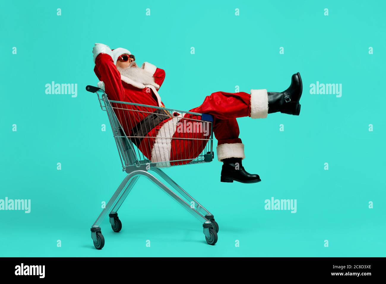 Vista laterale del simpatico Babbo Natale in occhiali da sole neri e in costume. Vecchio uomo che si diverte, sedendosi e rilassandosi nel carrello dello shopping sullo sfondo blu dello studio. Concetto di vacanza, divertimento. Foto Stock