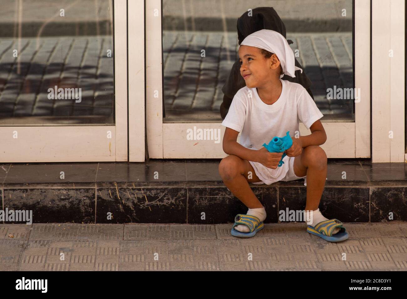 Havana / Cuba - 04.15.2015: Carino capretto cubano afro vestito di bianco seduto per strada, giocando con il suo giocattolo e sorridendo Foto Stock
