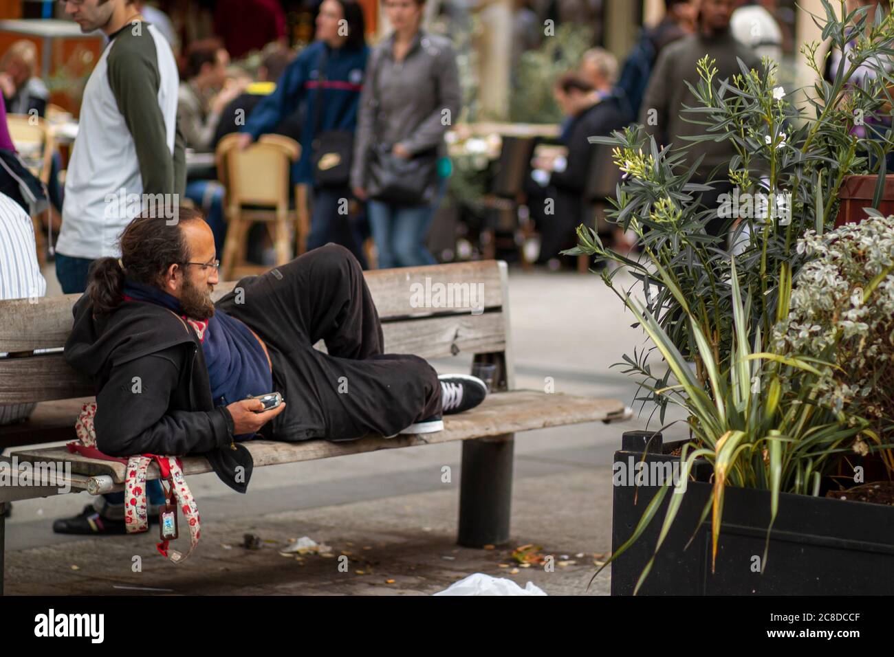 Parigi, Francia 06/12/2010: Un giovane senzatetto che indossa una felpa con cappuccio, sneakers e pantaloni da pista è sdraiato sul gomito su una panchina di legno. Sta controllando il suo Foto Stock