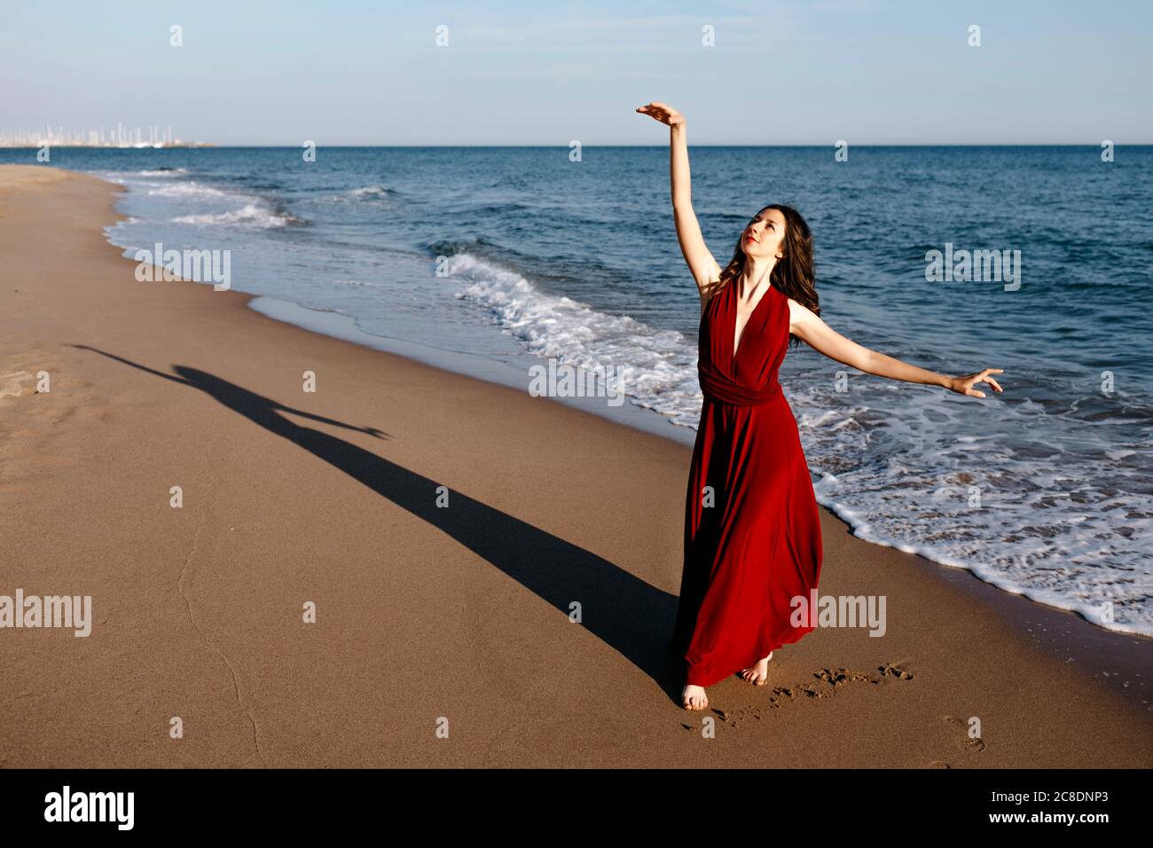 Donna delicata in abito rosso che danzano al mare, sentendo il sole Foto Stock