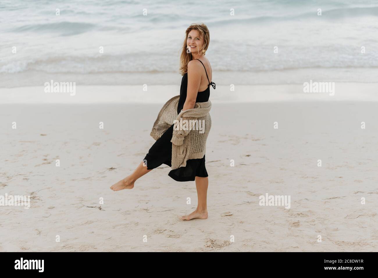 Donna mid adult sorridente che indossa un abito nero che cammina sulla spiaggia Foto Stock