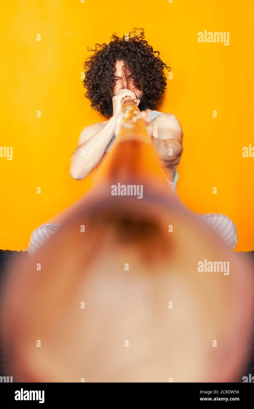 Uomo pazzo con capelli ricci che gioca didgeridoo mentre si siede contro sfondo giallo Foto Stock