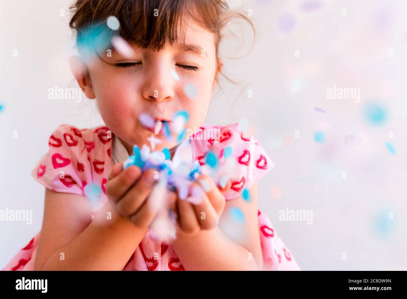 Primo piano di ragazza carina con gli occhi chiusi soffiando confetti da mani su sfondo bianco Foto Stock