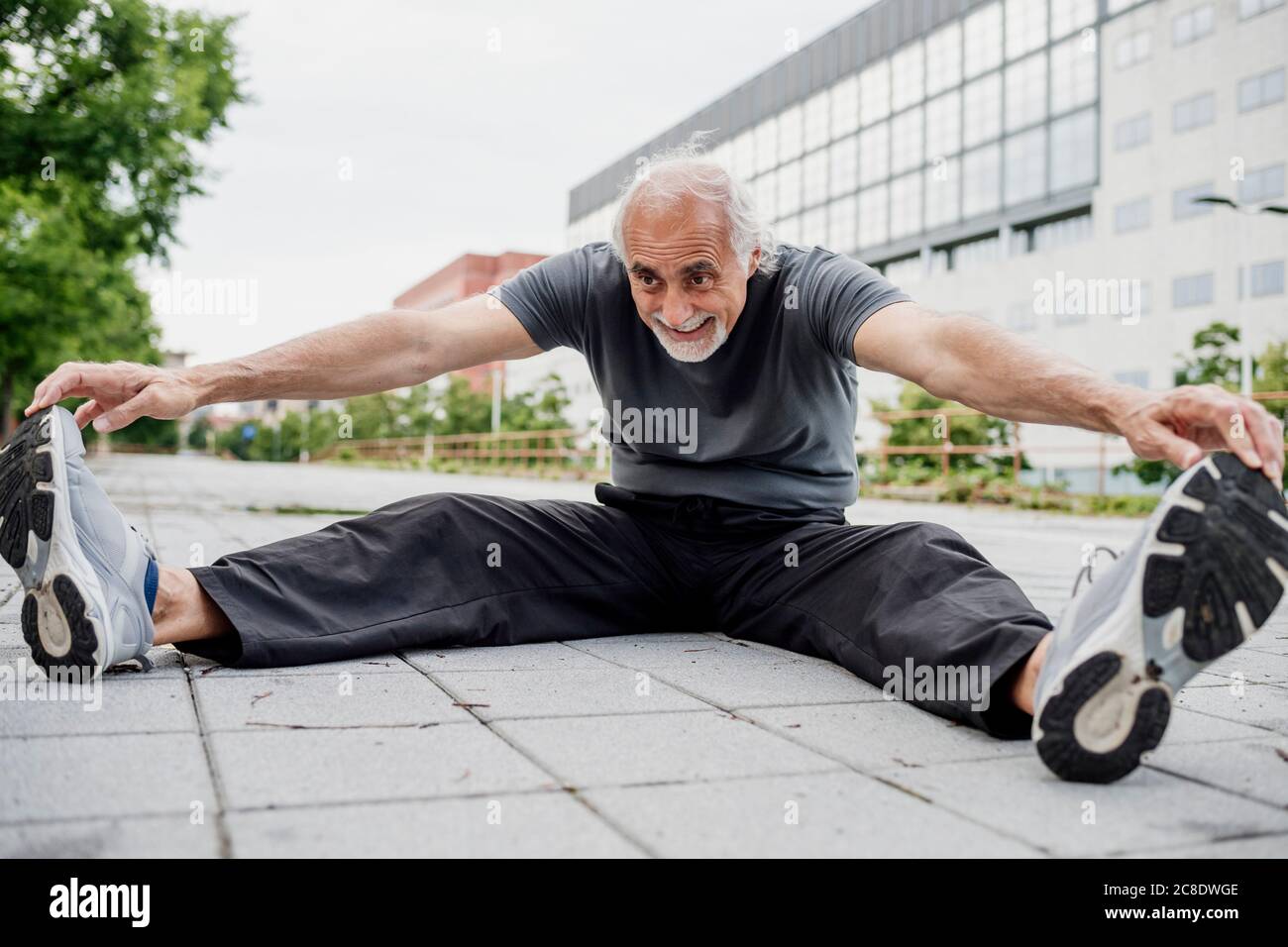 Sorridente uomo anziano che allunga il corpo mentre si siede sul sentiero dentro città Foto Stock