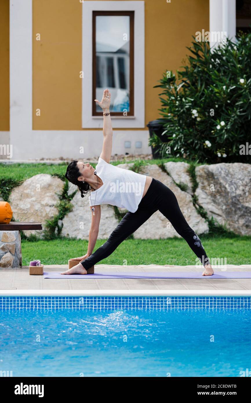 Donna che pratica posizione triangolo a bordo piscina contro casa in giardino Foto Stock