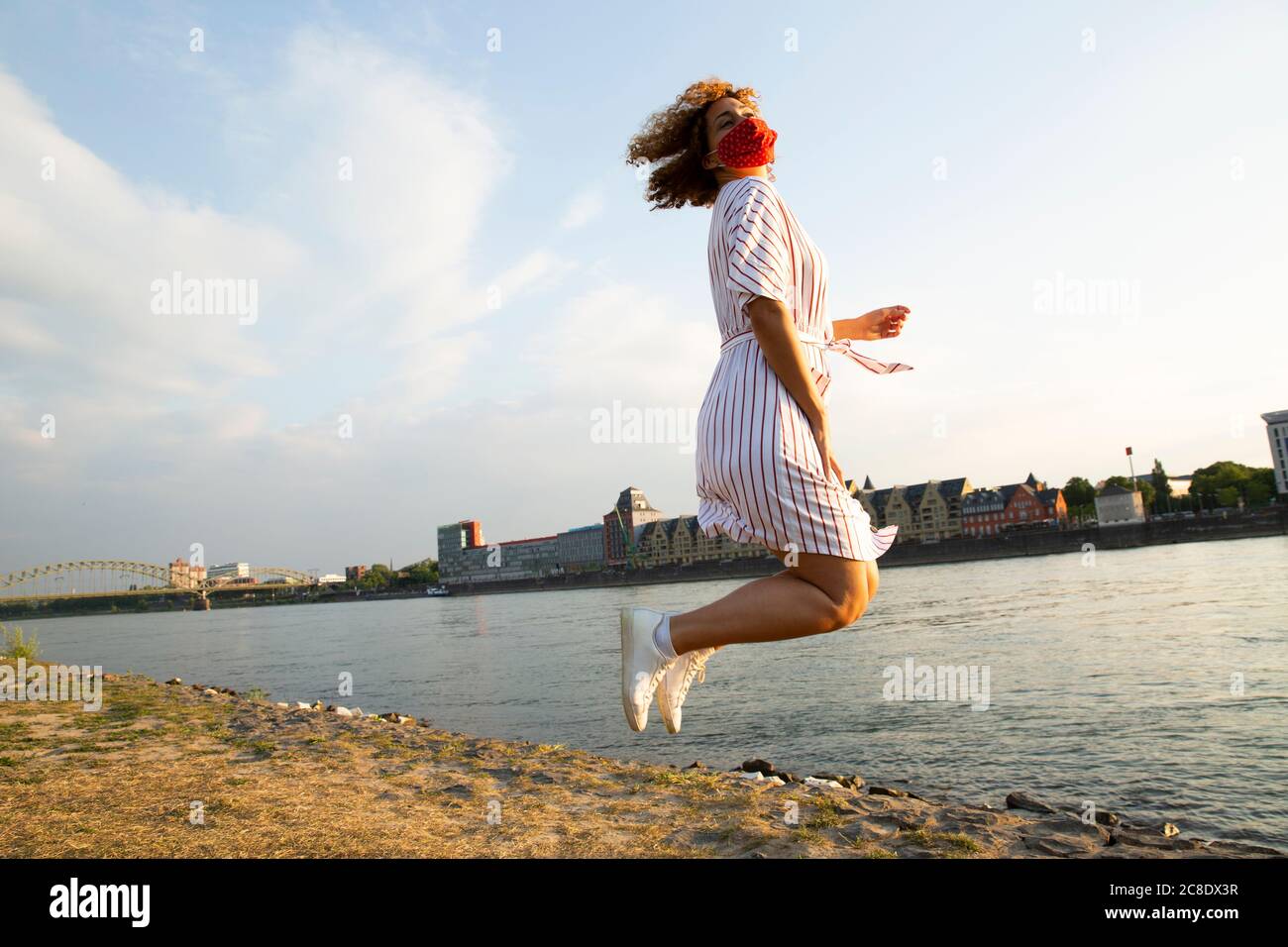 Donna mid adult che indossa una maschera che salta sulla riva del fiume contro il cielo in città Foto Stock