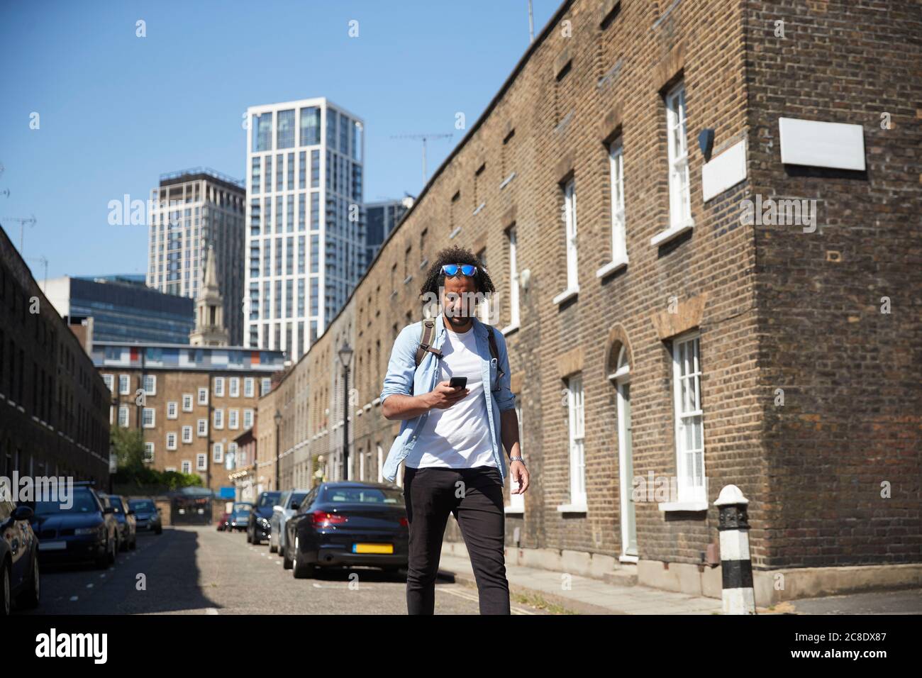 Giovane uomo che cammina su una strada residenziale guardando il cellulare, Londra, Regno Unito Foto Stock