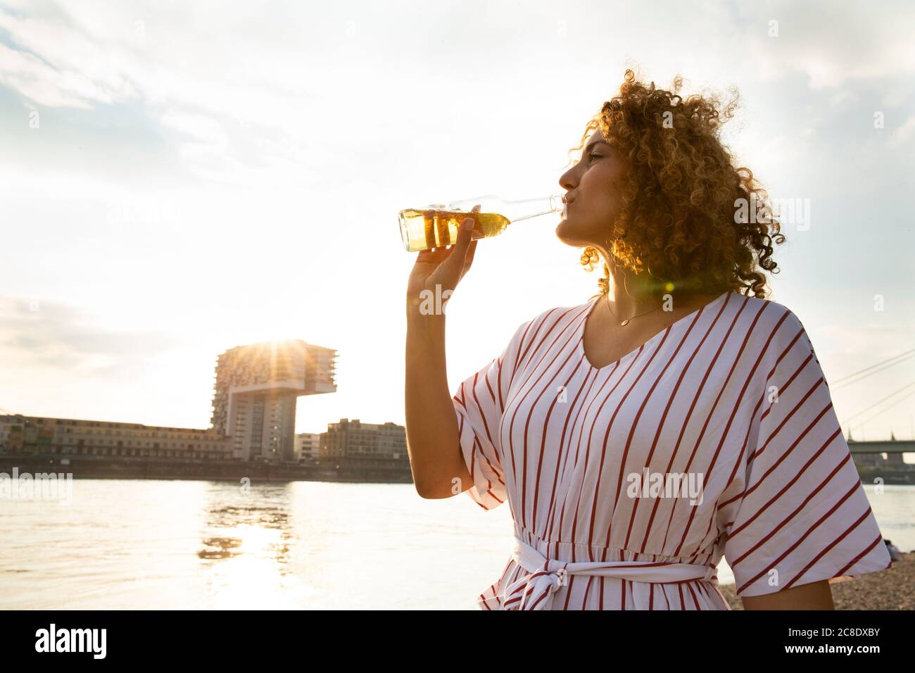 Donna mediamente adulta con capelli ricci che beve birra mentre si è in piedi contro il cielo in città al tramonto Foto Stock