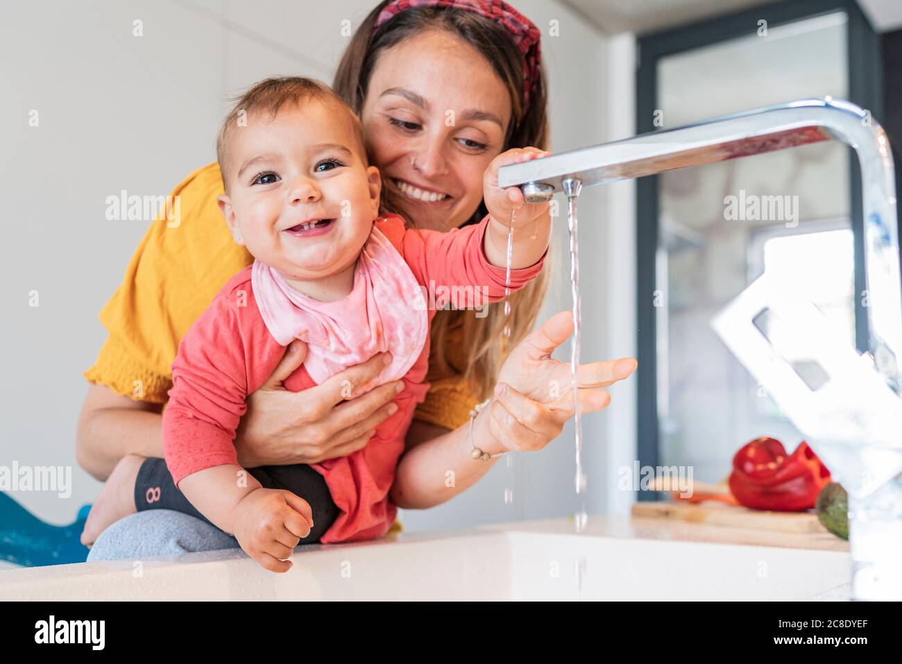 Madre sorridente e bambina simpatica che gioca con l'acqua che cade dal rubinetto nel lavandino della cucina Foto Stock