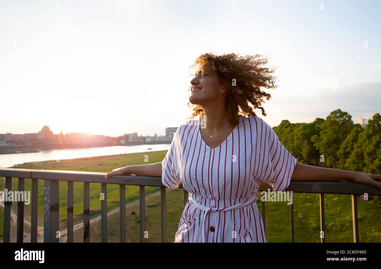 Donna mid adulta sorridente con capelli ricci in piedi dalla ringhiera contro il cielo al tramonto Foto Stock