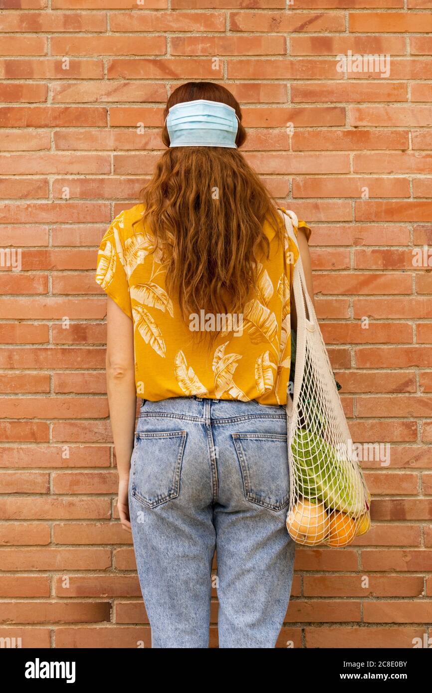 Giovane donna con borsa della spesa della spesa che indossa la maschera facciale capelli mentre in piedi davanti a muro di mattoni Foto Stock