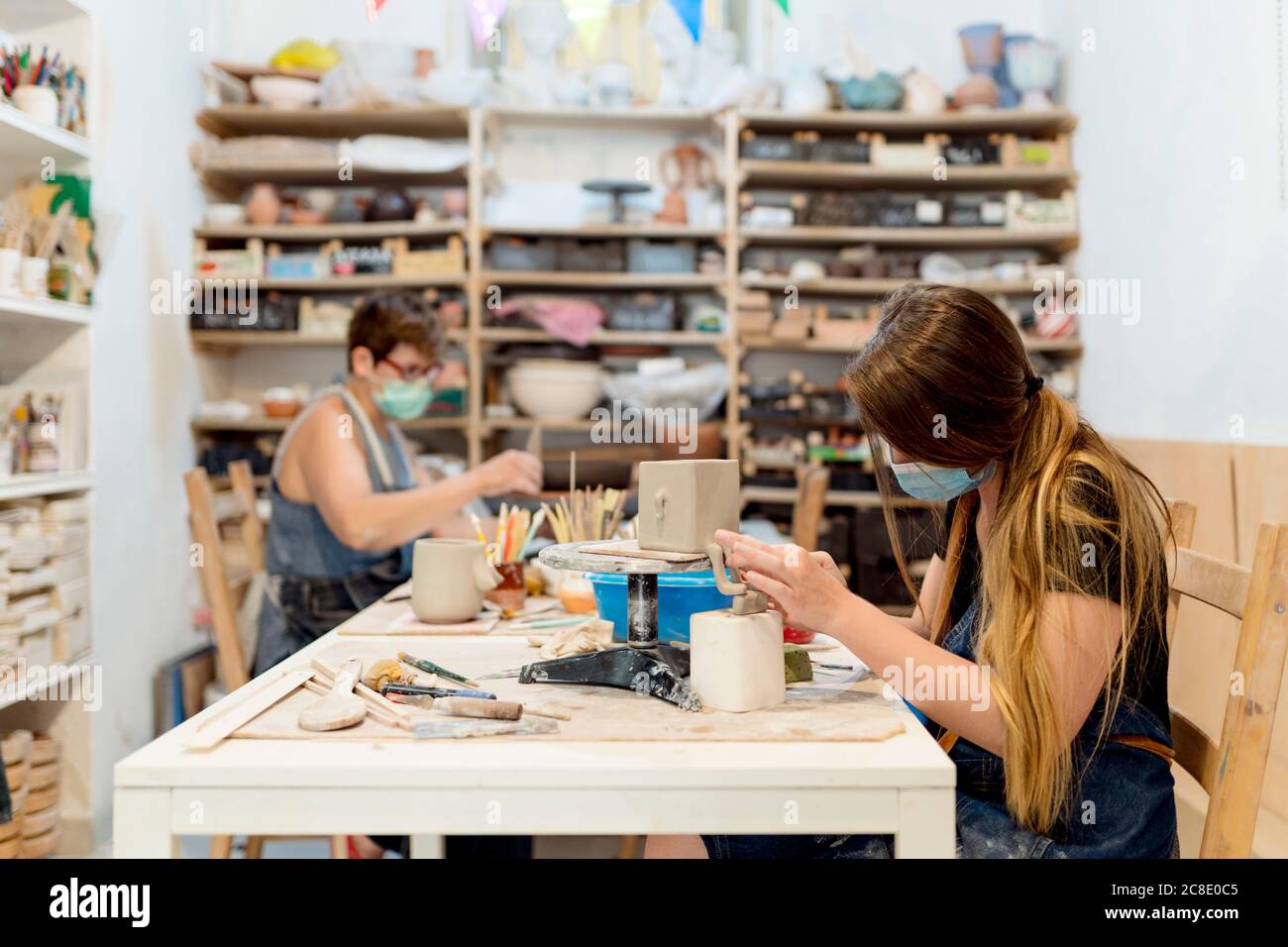 Donne che indossano maschere che fanno ceramica sul banco in officina Foto Stock