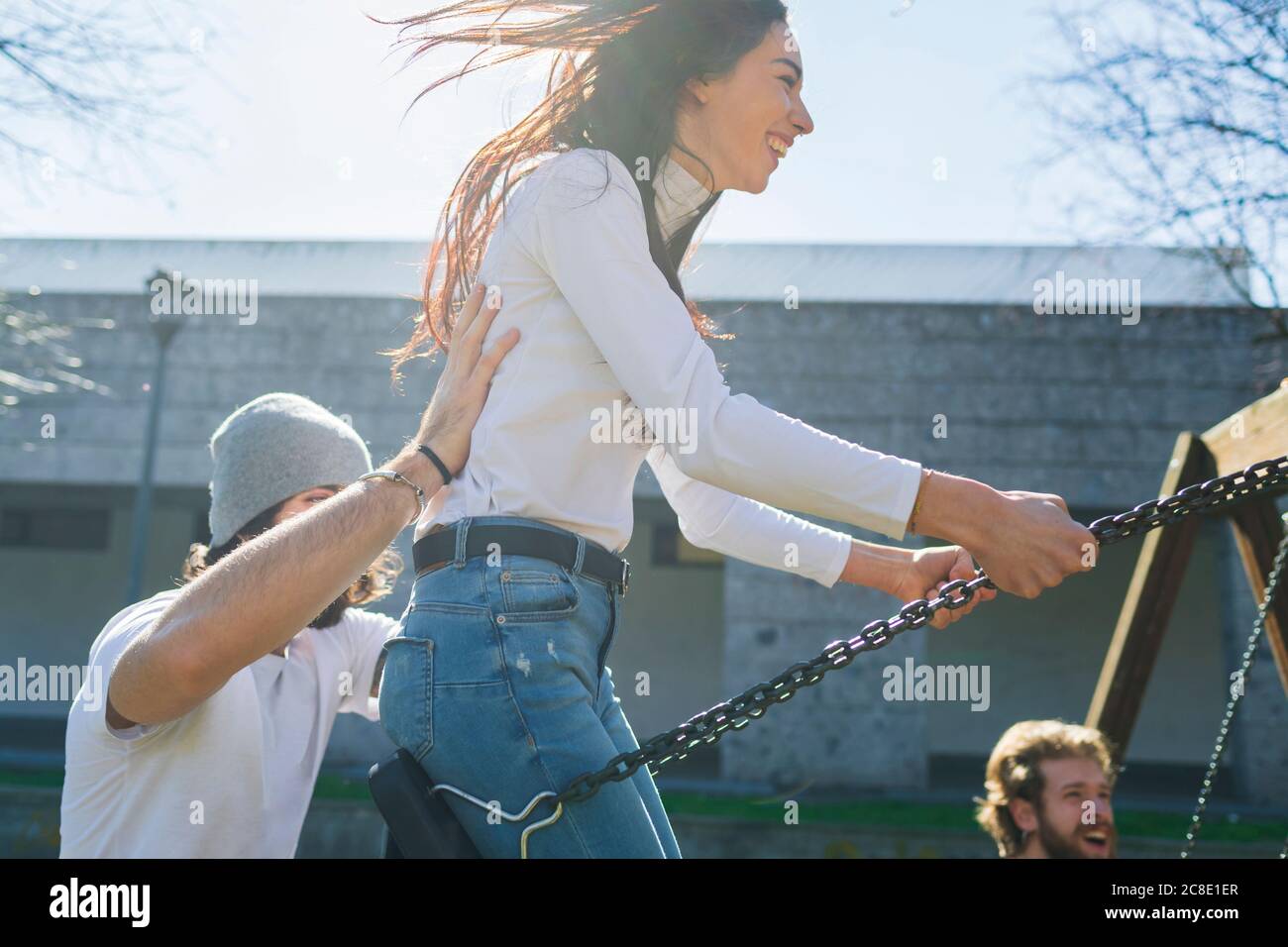 Giovane uomo che spinge amici giocosi che oscillano nel parco durante il sole giorno Foto Stock