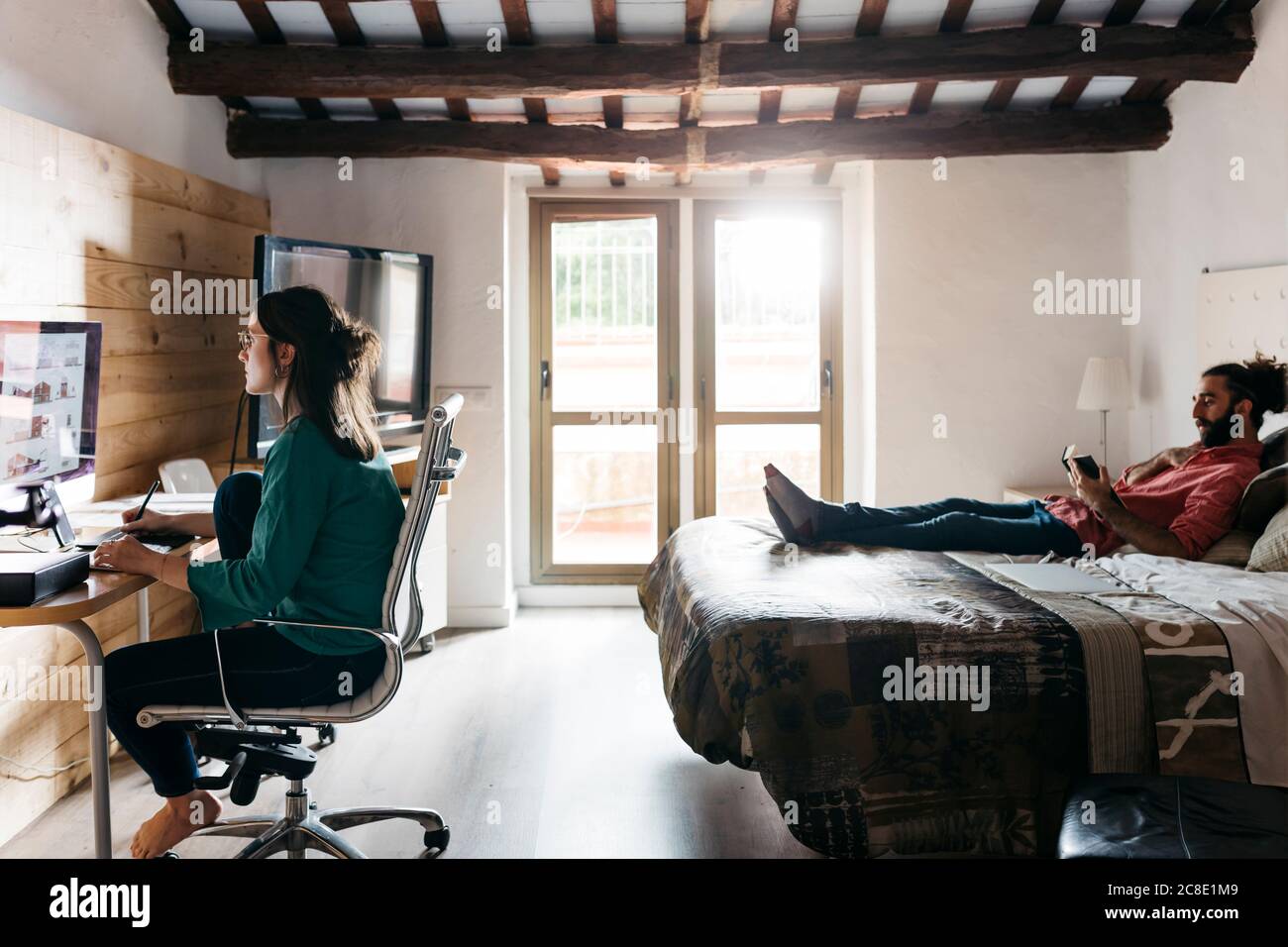 Architetto femminile che usa il computer a casa, uomo che legge un libro sdraiato sul letto Foto Stock