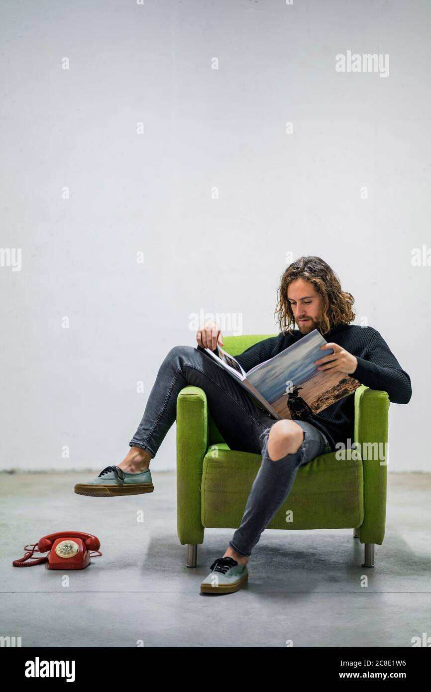 Giovane uomo che legge libro mentre si siede sulla poltrona contro il muro a casa Foto Stock