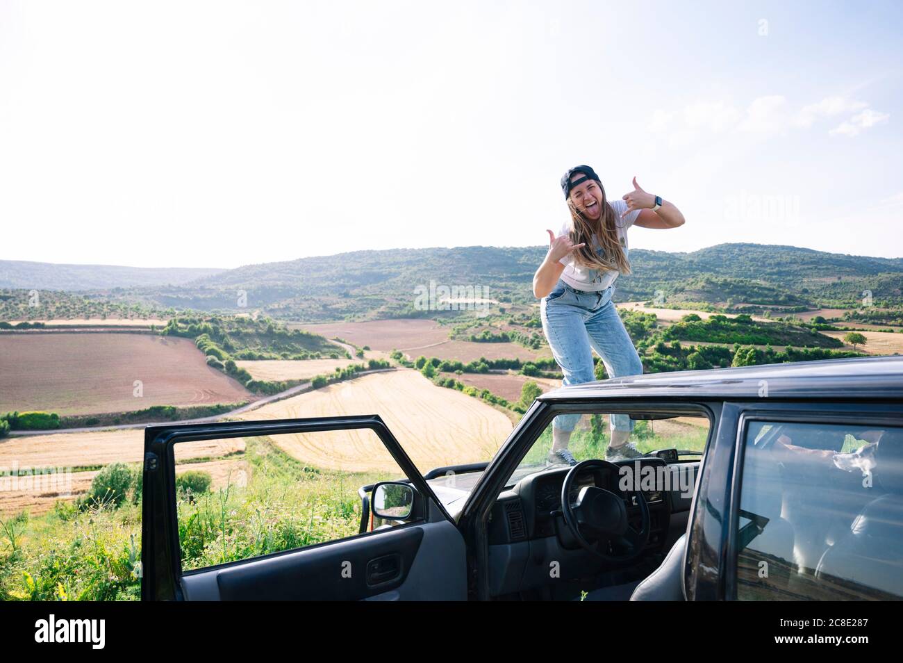 Donna allegra che si stacca la lingua mentre mostra il segno della mano sopra cofano del veicolo contro il cielo Foto Stock