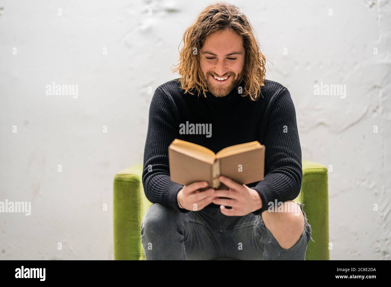 Uomo sorridente che legge libro mentre si siede sulla poltrona contro il muro a casa Foto Stock