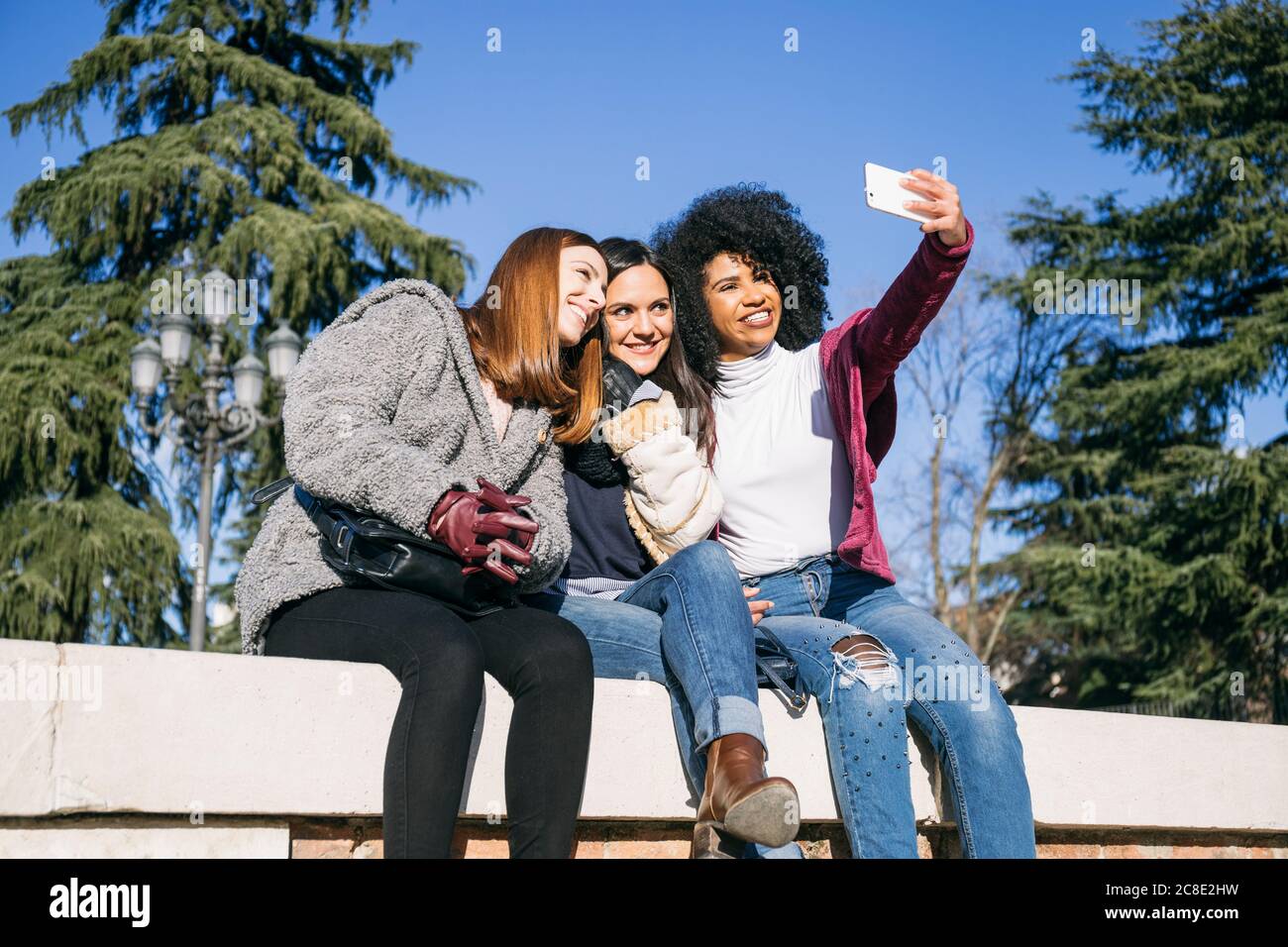 Donna che prende selfie con le amiche felici mentre si siede sopra parete di ritegno in posizione di parcheggio Foto Stock