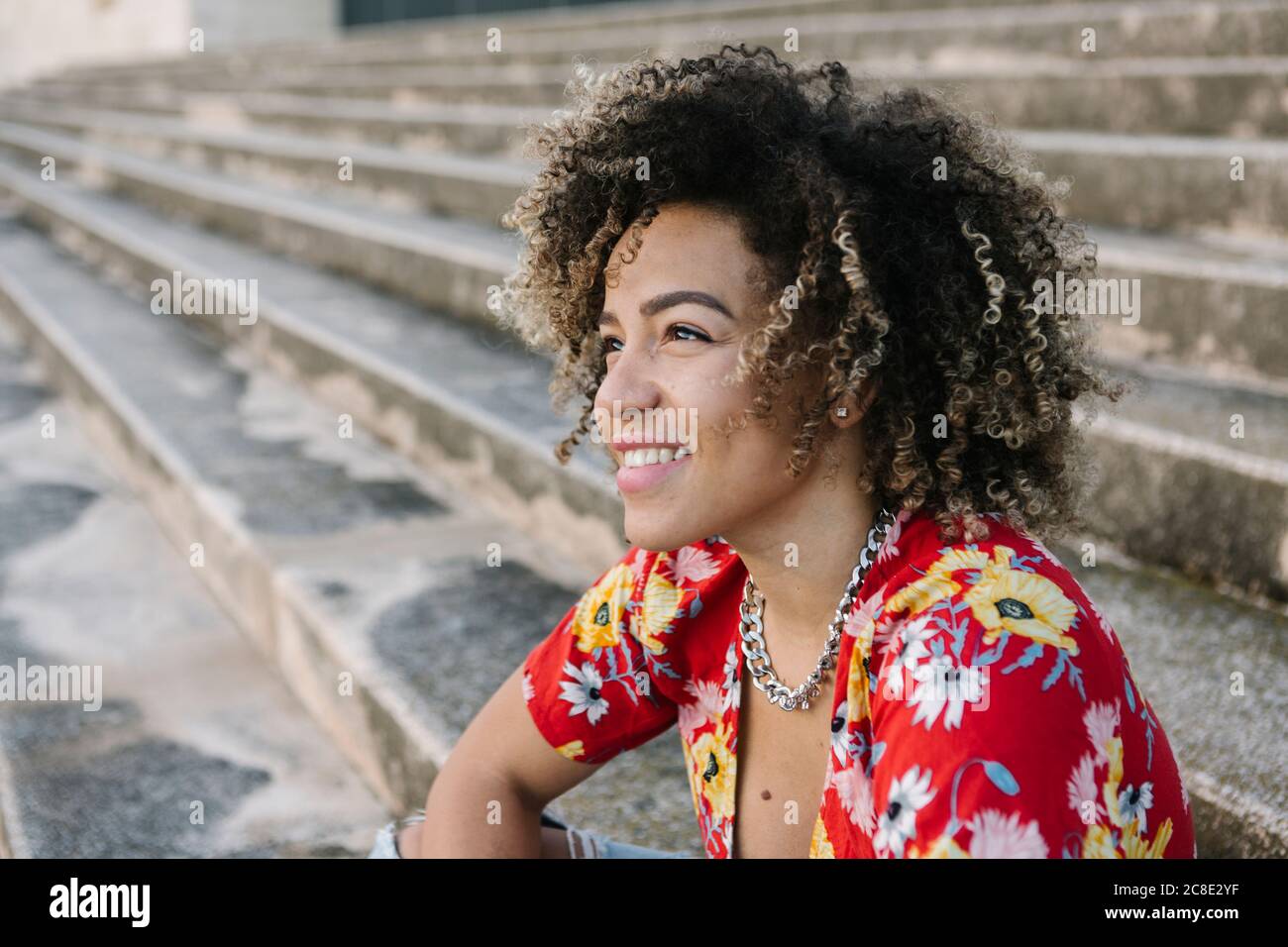 Giovane donna premurosa sorridente mentre si siede sui gradini durante il sole giorno Foto Stock