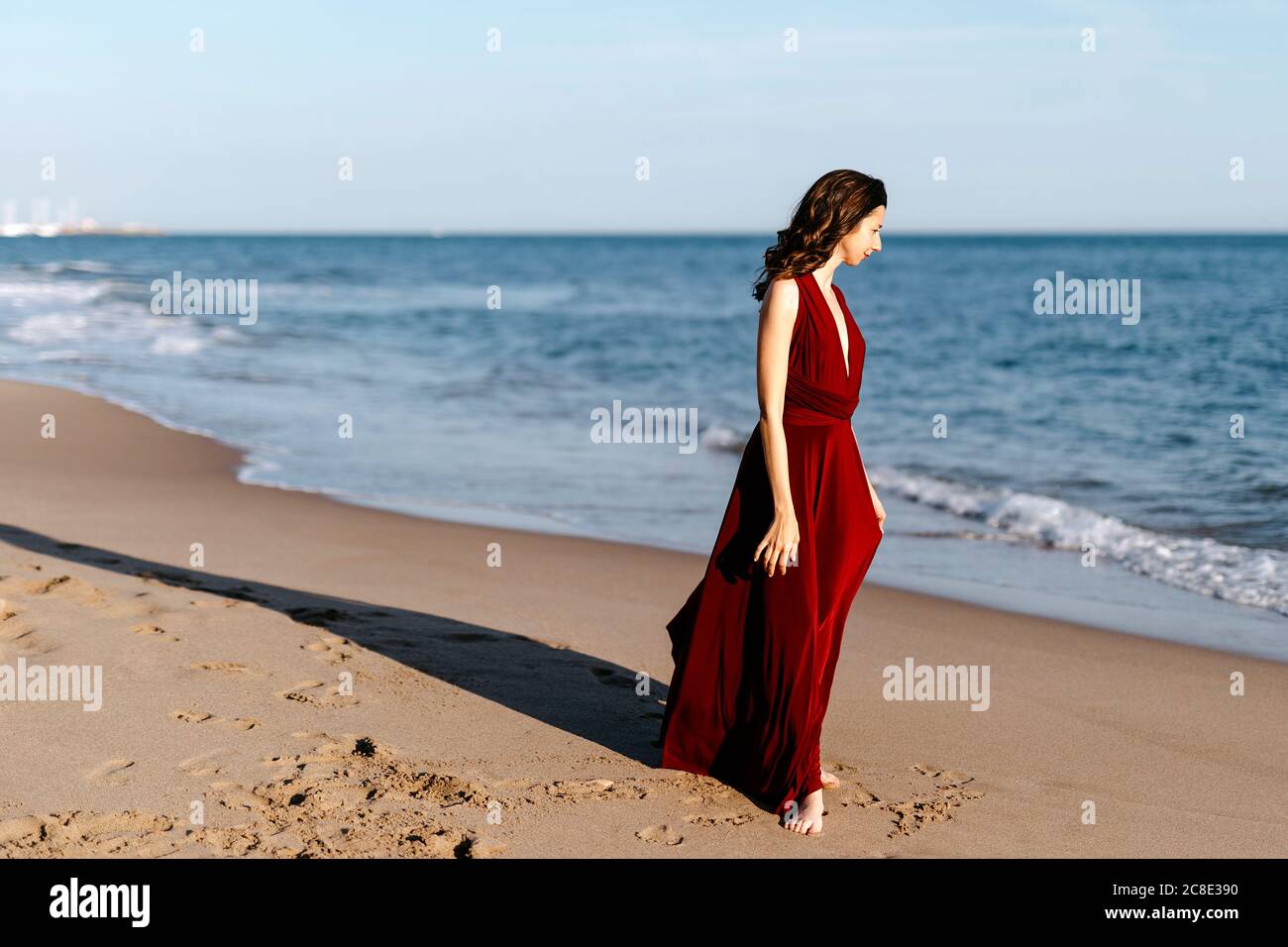 Donna delicata in abito rosso che cammina al mare, sentendo il sole Foto Stock