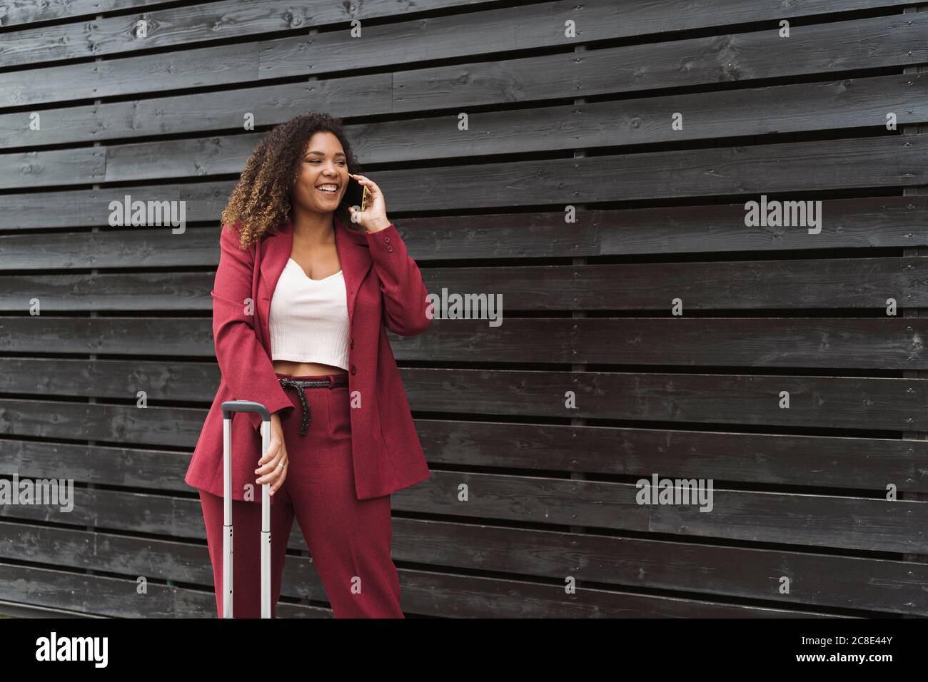 Sorridente bella imprenditore femminile che parla sul telefono cellulare mentre si è in piedi con bagagli contro muro di legno nero Foto Stock