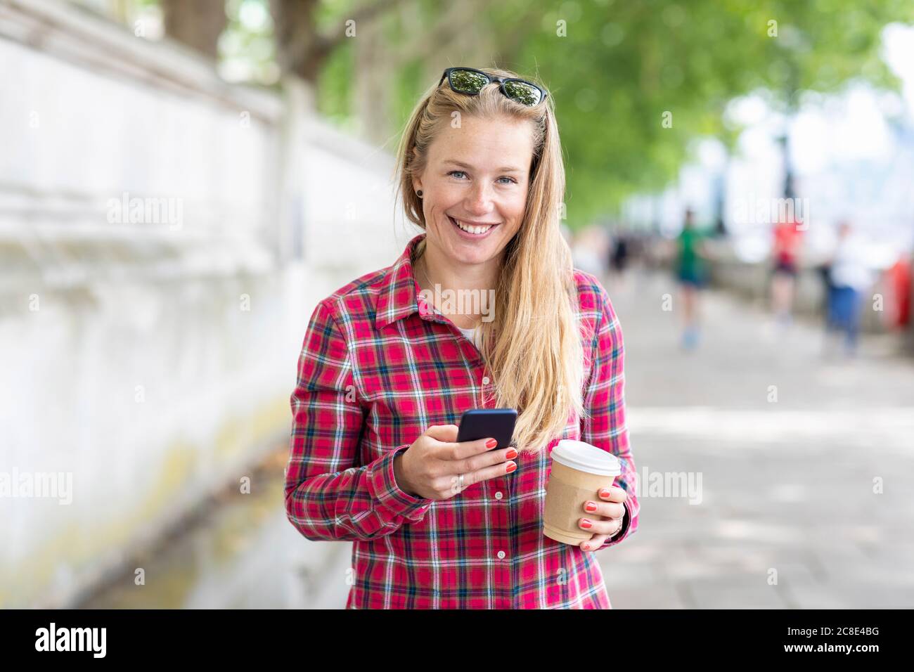 Donna sorridente che tiene il caffè utilizzando il telefono cellulare mentre si sta in piedi sentiero Foto Stock