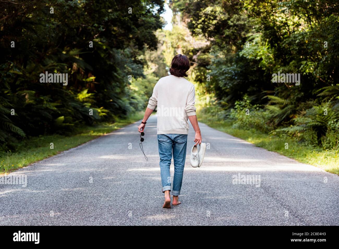 Uomo che tiene scarpe sportive e occhiali da sole mentre cammina su strada tra alberi Foto Stock