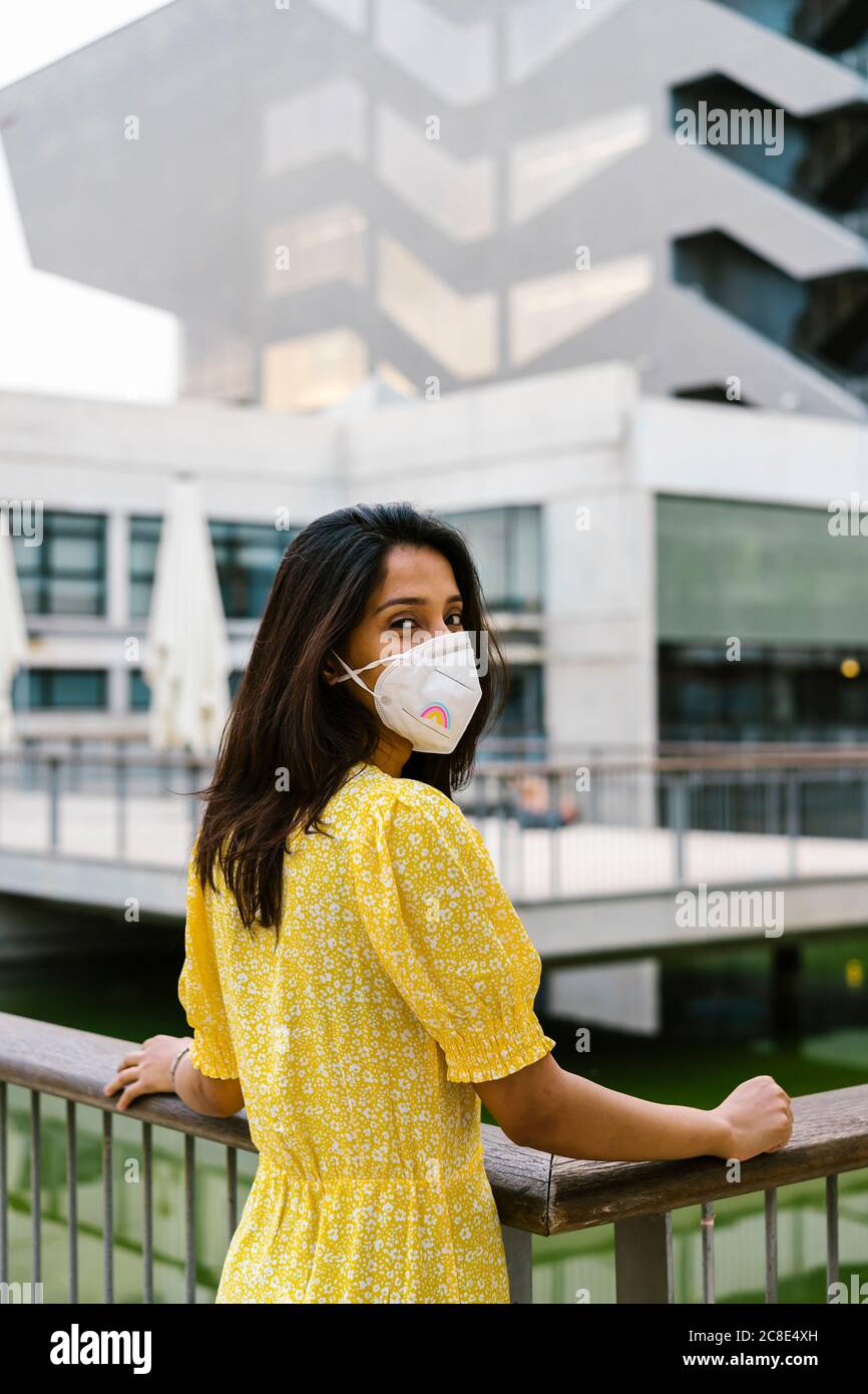 Giovane donna che indossa la maschera mentre si trova su una passerella sopraelevata in città Foto Stock