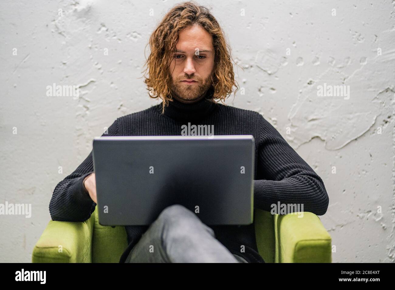 Ragazzo sopportato usando il laptop mentre si siede sulla poltrona contro parete a casa Foto Stock