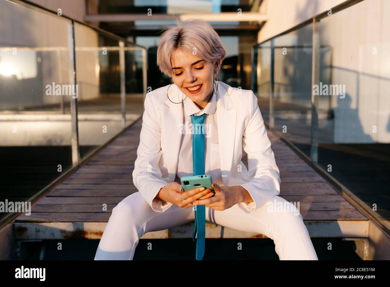 Giovane donna che indossa un vestito bianco con smartphone mentre si è seduti contro un edificio moderno Foto Stock