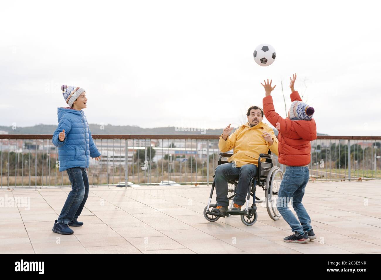 Uomo seduto su sedia a rotelle che lancia la palla verso i figli giocosi dentro parcheggio Foto Stock