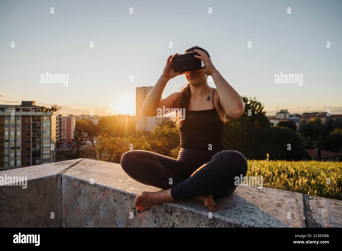 Donna sportiva che usa gli occhiali VR mentre si siede sulla parete di ritegno in città durante il tramonto Foto Stock