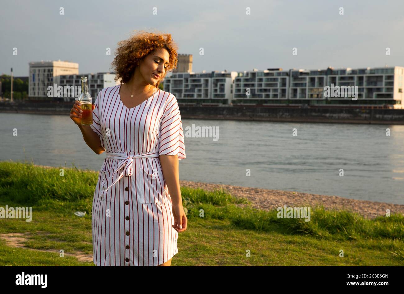 Donna mid adult che tiene una bottiglia di birra in piedi su terra erbosa contro il fiume in città al tramonto Foto Stock
