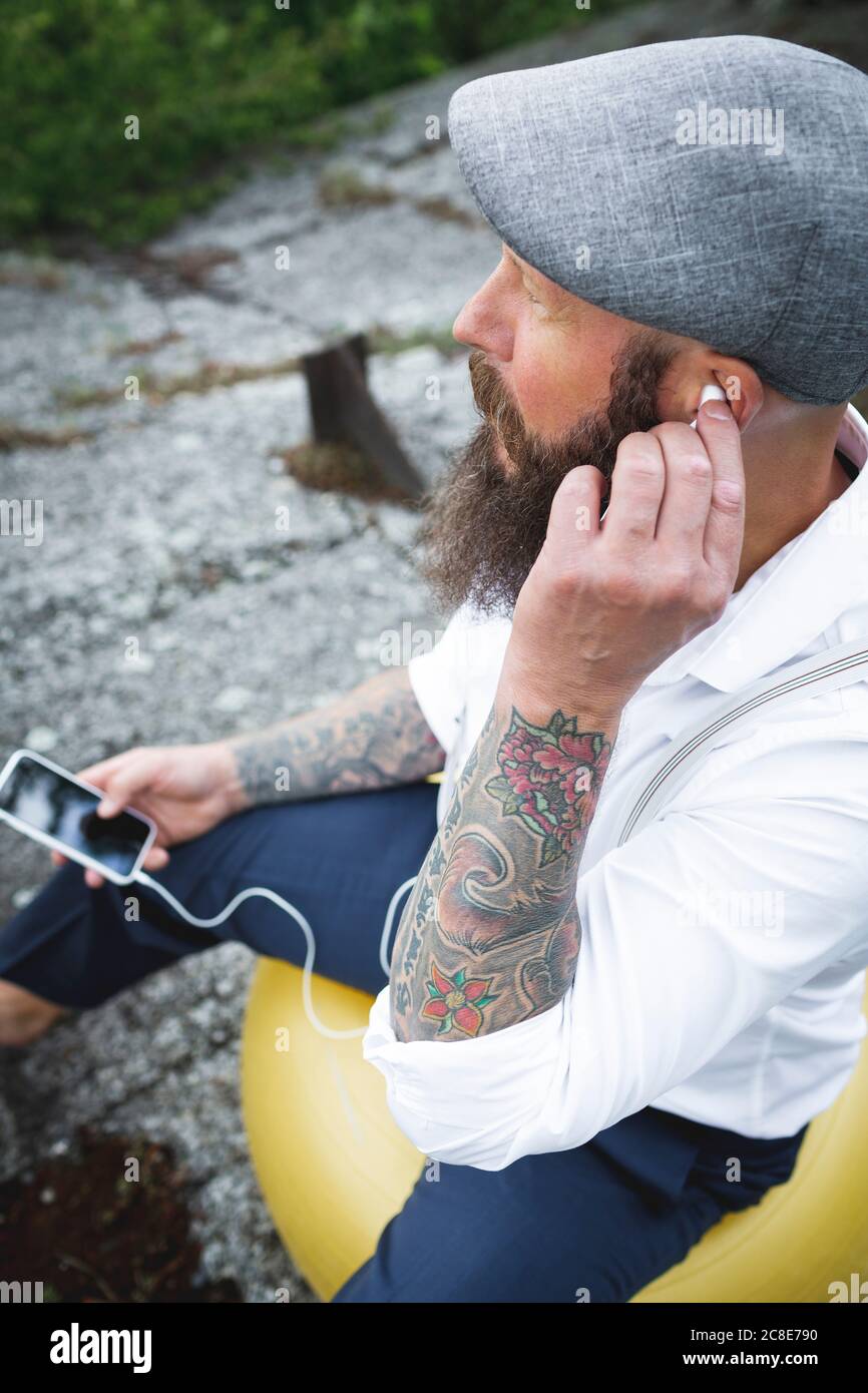 Primo piano di un uomo con cappuccio grigio che ascolta la musica attraverso le cuffie mentre si siede sulla palla da fitness Foto Stock