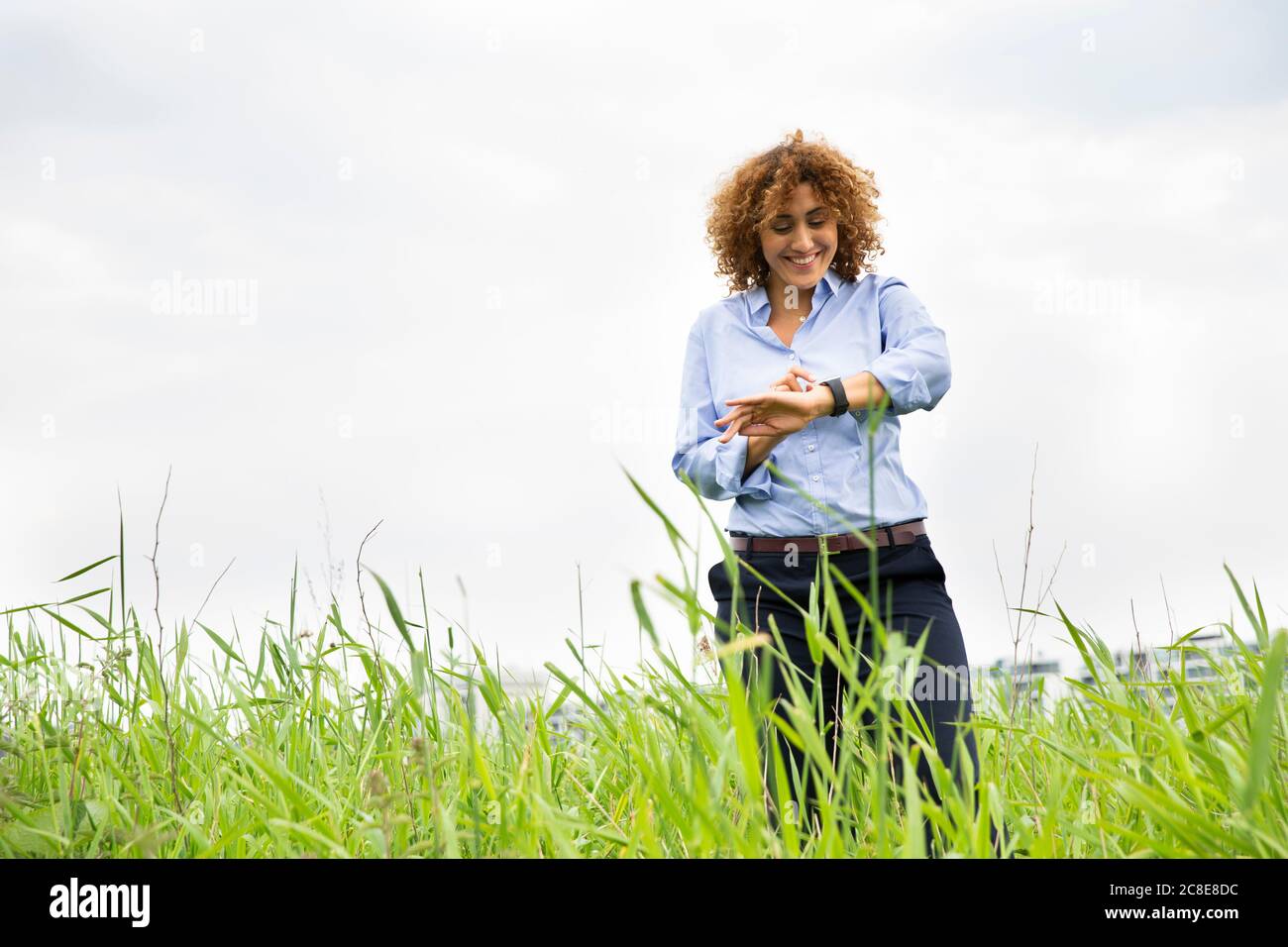 Donna d'affari sorridente che controlla il tempo mentre si sta in piedi su terra erbosa contro cielo nuvoloso Foto Stock