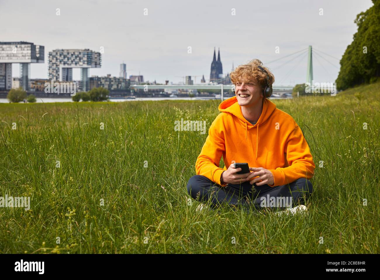 Felice giovane seduto su un prato ascoltando musica con le cuffie, Colonia, Germania Foto Stock