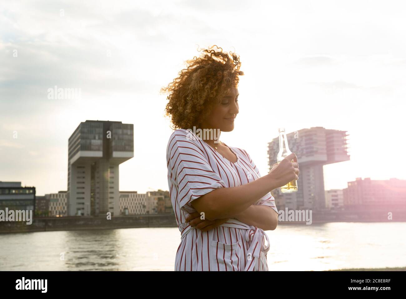 Donna mid adult che tiene una bottiglia di birra mentre si trova contro il fiume ed edifici in città Foto Stock