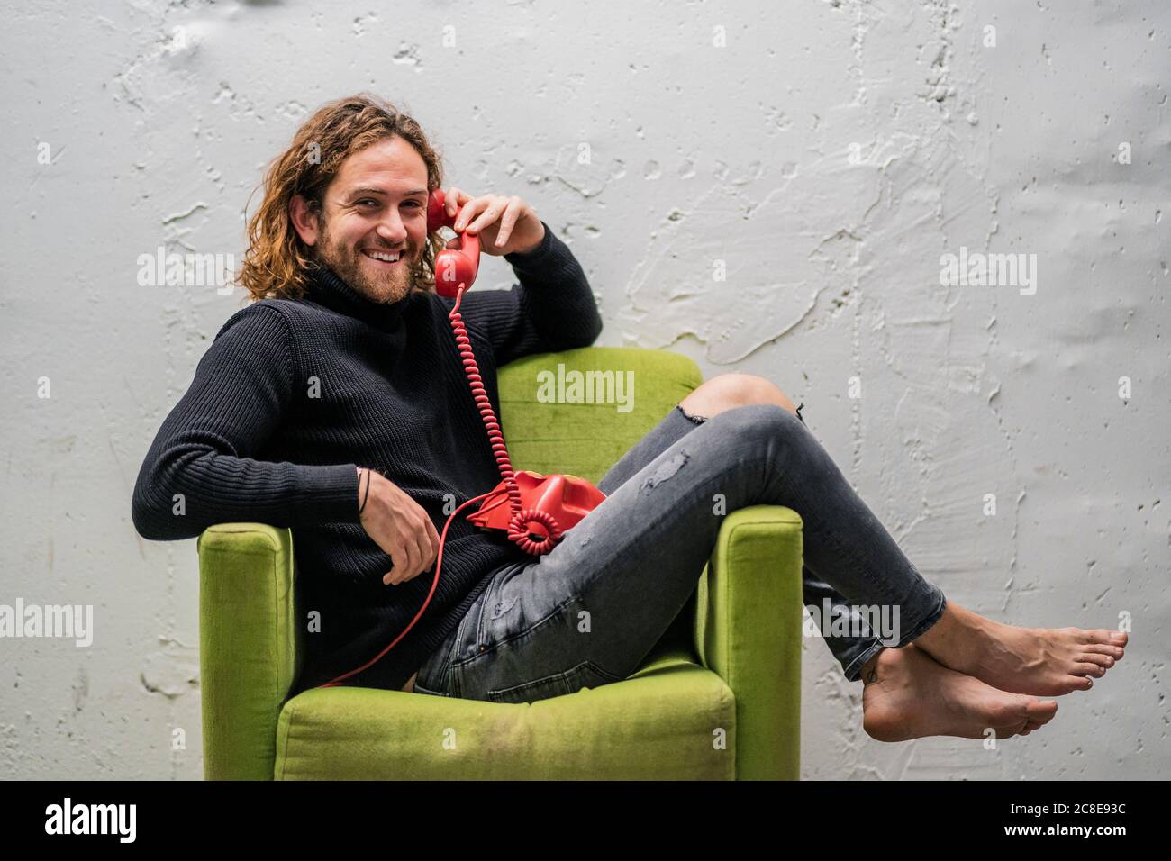 Uomo sorridente che parla al telefono mentre si rilassa sulla poltrona contro parete Foto Stock