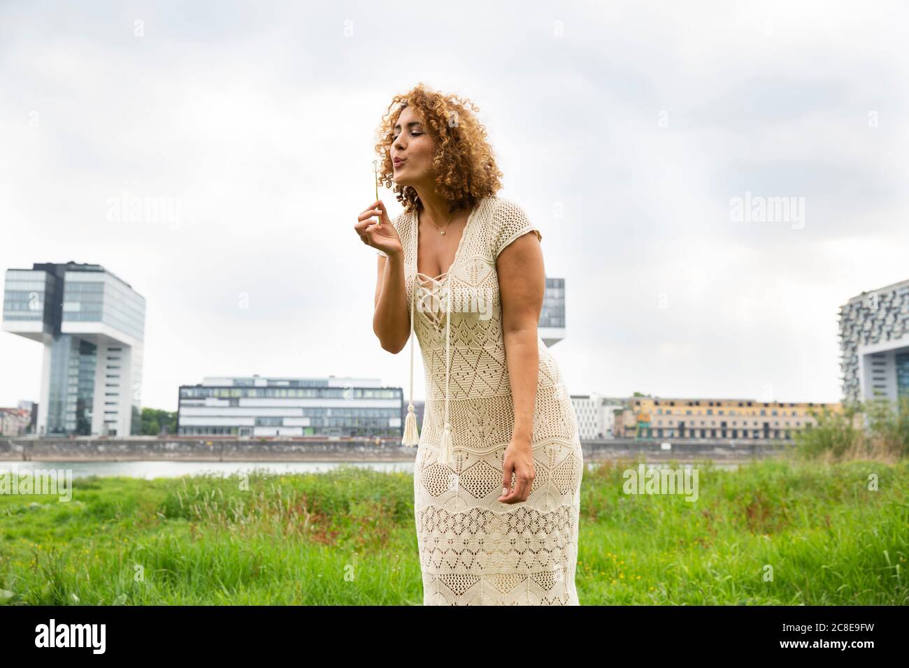 Donna mediamente adulta con capelli ricci che soffiano fiore mentre in piedi su terra erbosa contro il cielo Foto Stock