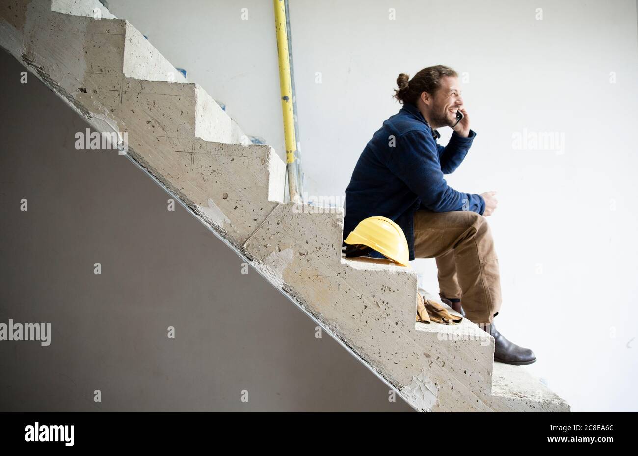 Il lavoratore della costruzione parla con lo smartphone mentre si siede sui gradini in costruzione di casa Foto Stock